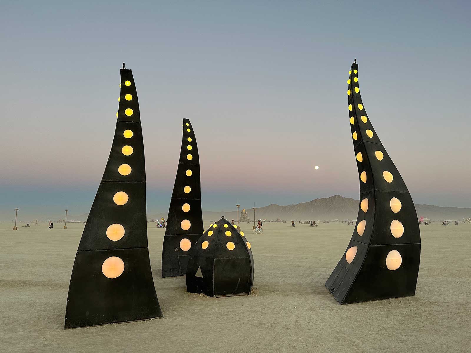 Three tall black spire sculptures with glowing dots at dusk moonrise