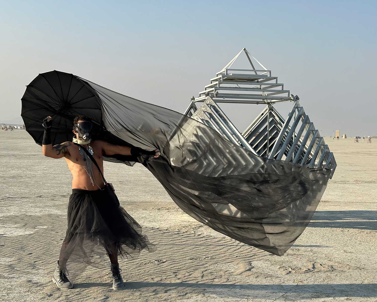 Masked participant in black flowing fabric poses near pyramid sculpture