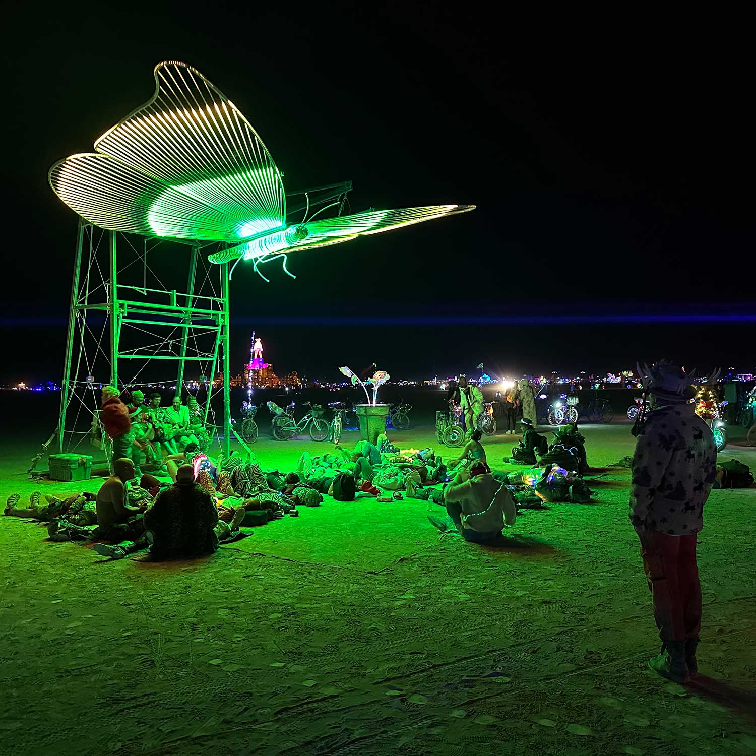 Green glowing dragonfly sculpture above resting crowd on the playa