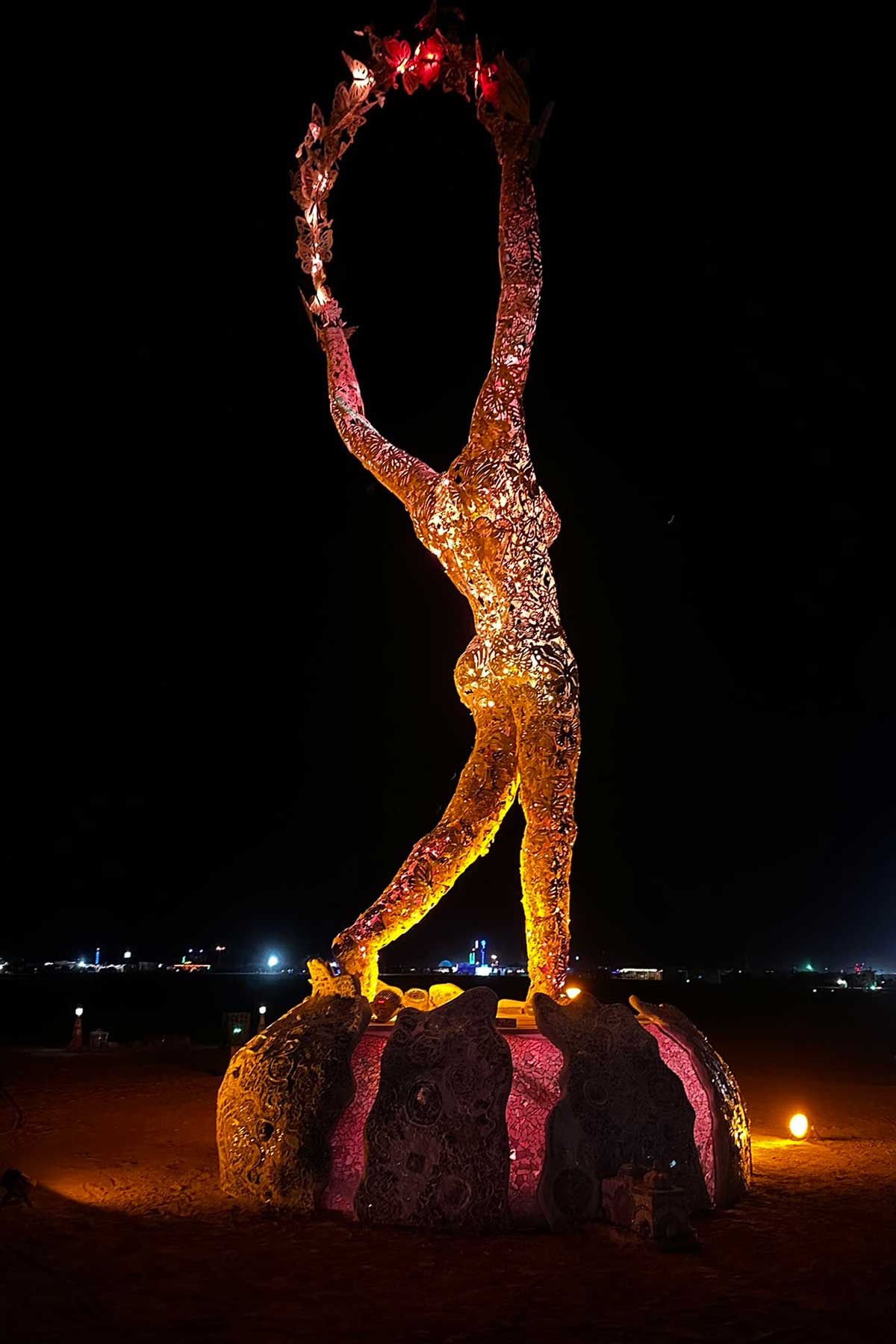 Tall illuminated woman figure holding floral hoop aloft at night