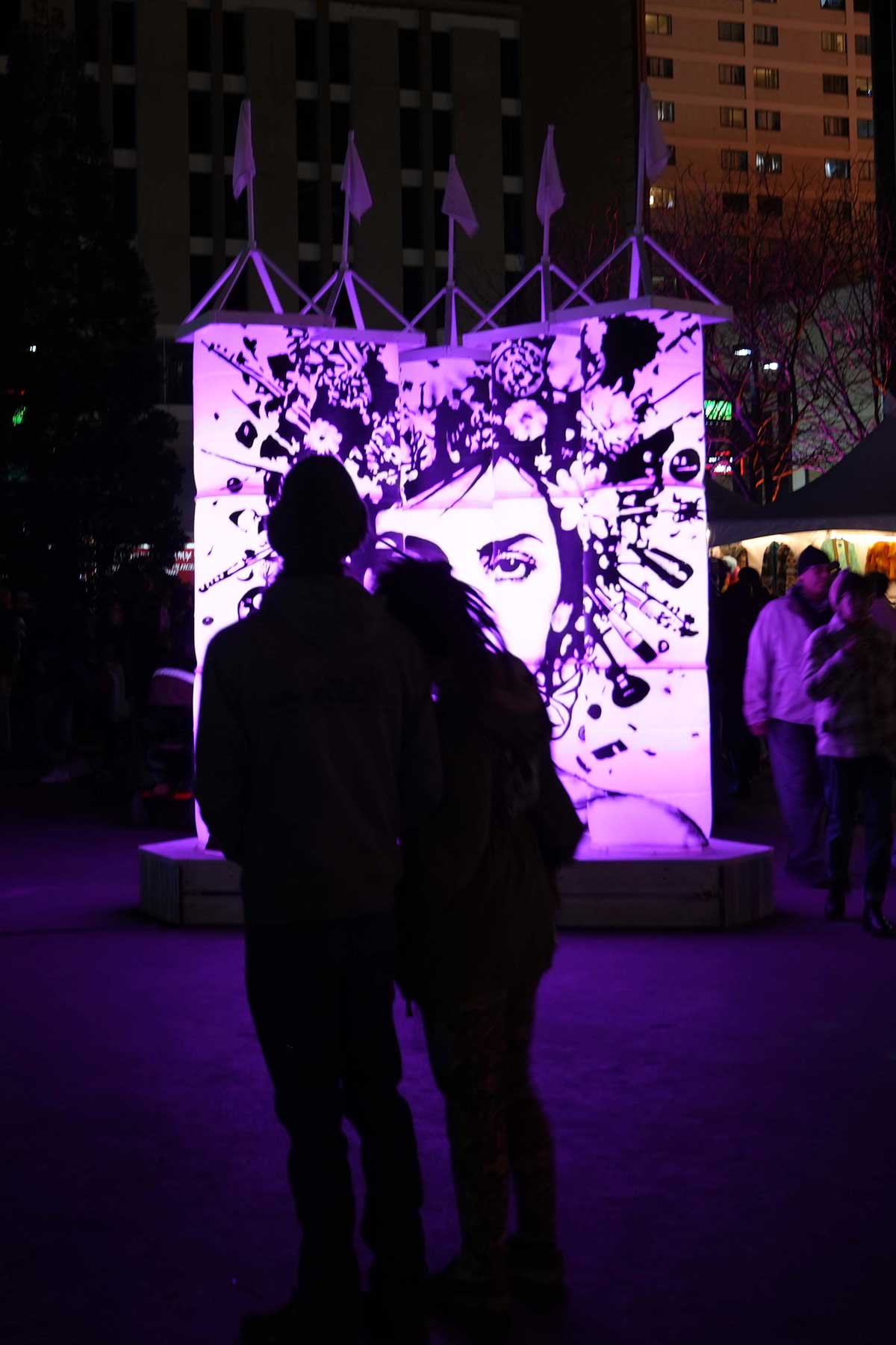 Couple silhouetted before large purple-lit face art installation at night