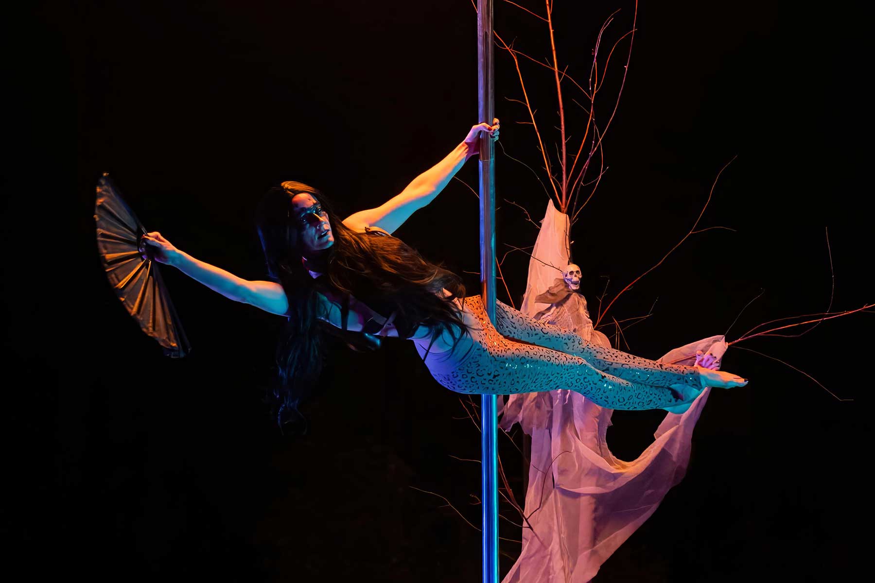 Pole dancer in mermaid costume holds fan beside skull prop