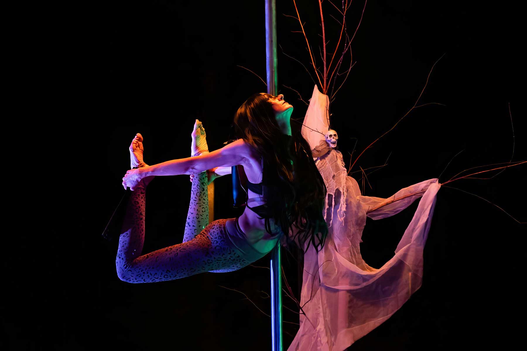 Leopard-print pole dancer stretched horizontal on pole beside skull prop