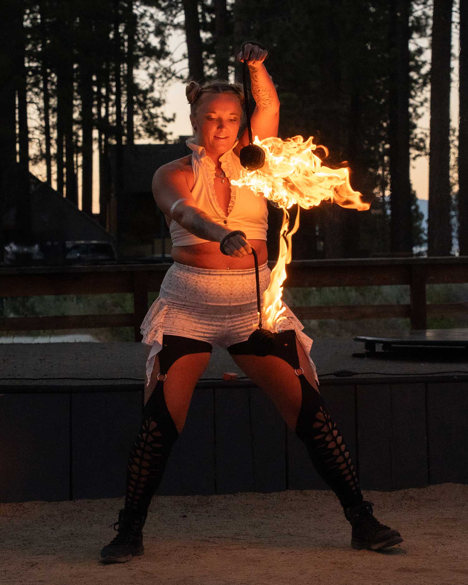 Fire performer spinning flaming poi outdoors against sunset pine trees