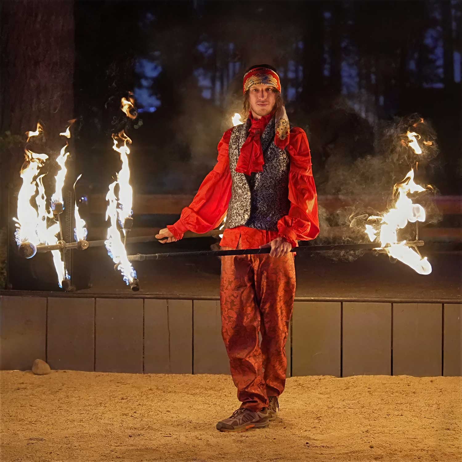 Costumed performer holding fire staff horizontally at sandy outdoor venue