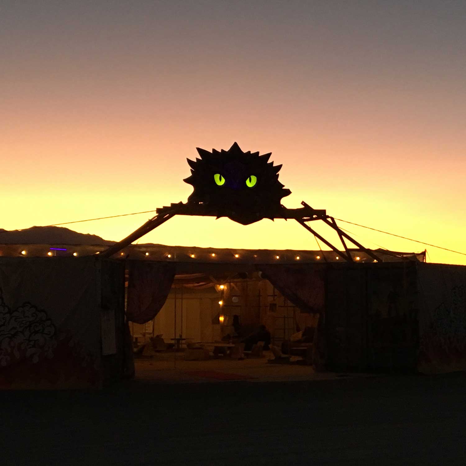 Burning Man camp entrance with glowing green-eyed creature at sunset