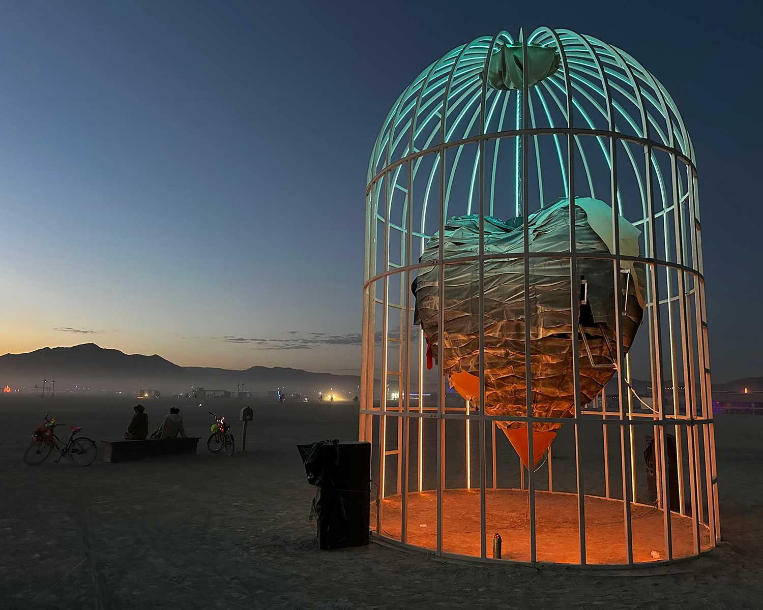 Giant illuminated birdcage sculpture on desert playa at sunset